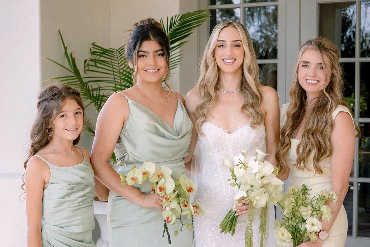 Bride with her bridesmaids in sage green and pale yellow gowns, holding white and green bouquets.