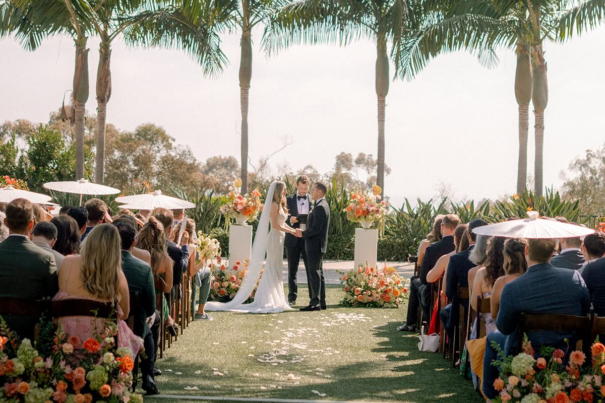 Outdoor ceremony on a palm-lined lawn beneath string lights, with guests seated along a flower-lined aisle.