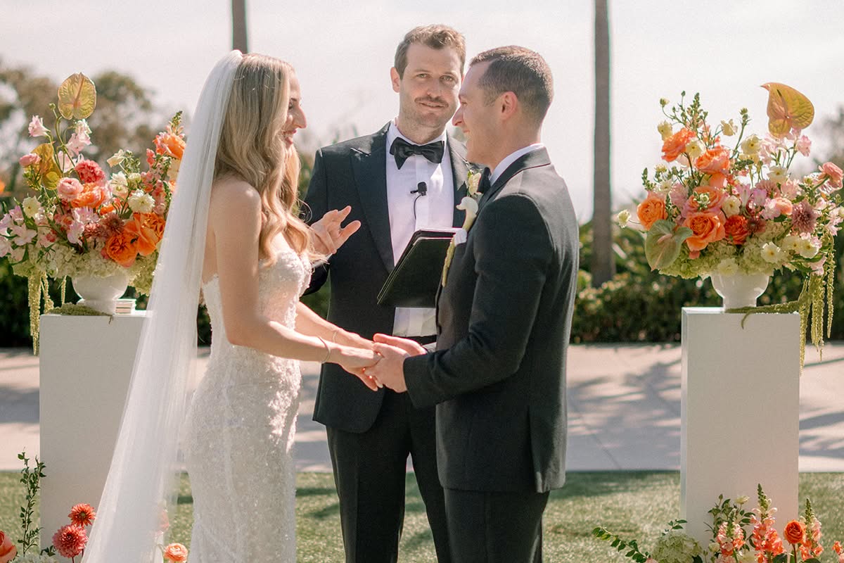 Bride and groom holding hands as they exchange vows between white pedestals topped with peach and coral florals.