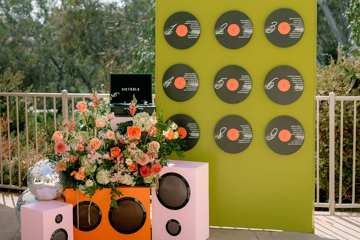 “Find your seat & drop a beat” seating chart made of vinyl records, with a record player, colorful speaker boxes, disco balls, and florals.
