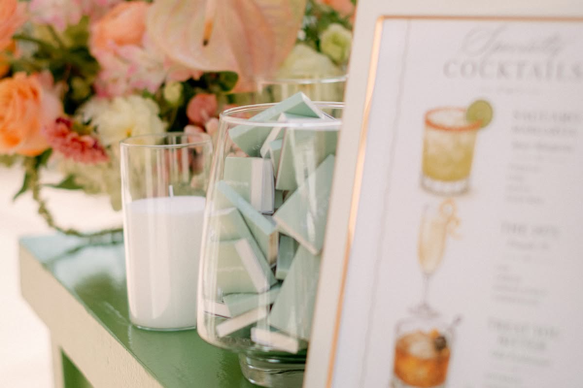Illustrated specialty cocktails sign on a green bar beside peach and coral florals.