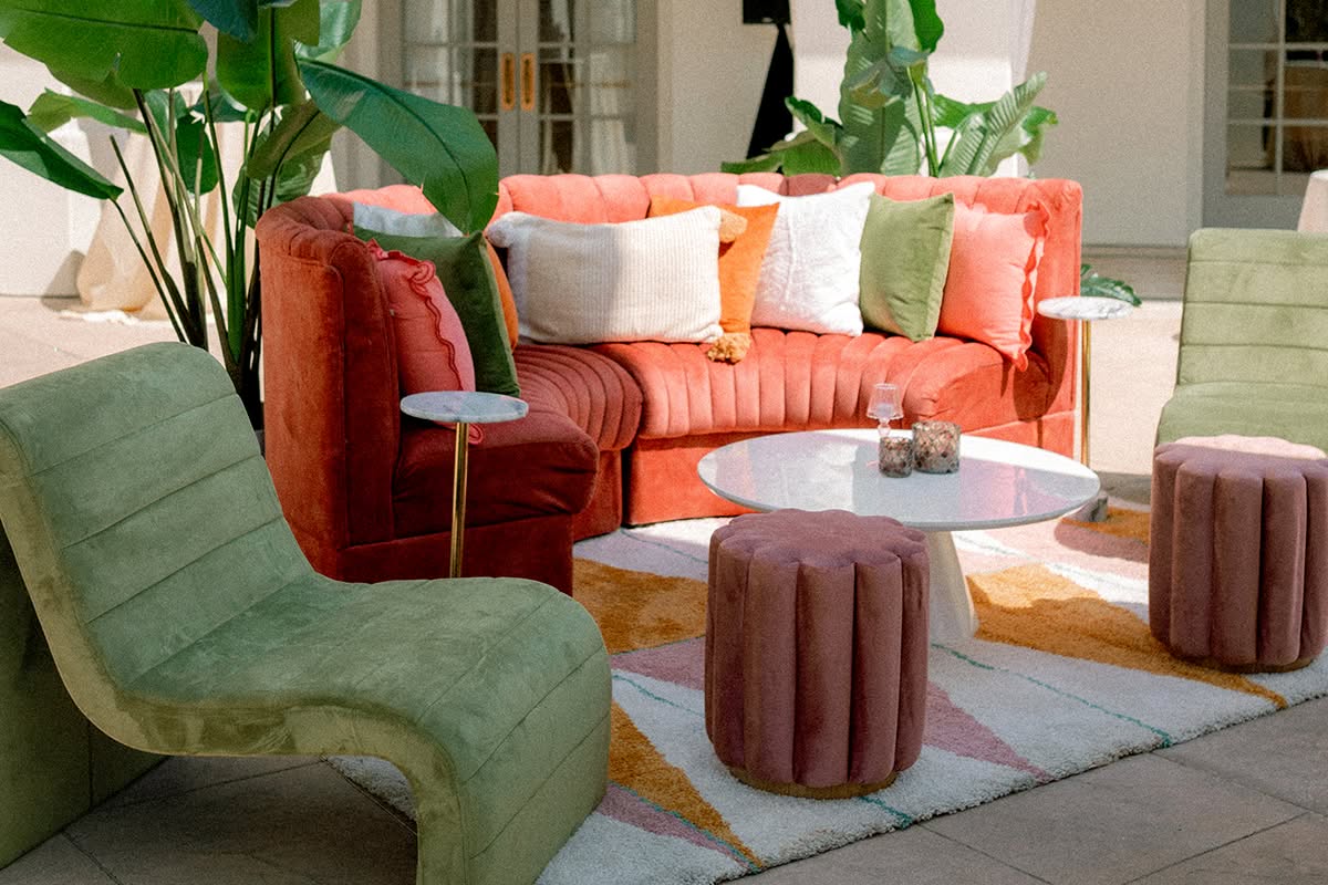 Courtyard lounge with a coral velvet sofa, sage green chairs, mauve ottomans, and a patterned rug.