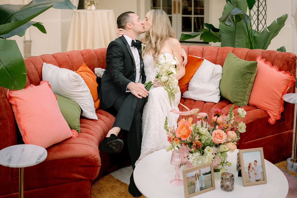 Alex and Cory sharing a kiss on a coral velvet sofa beside a table of florals and framed photos.