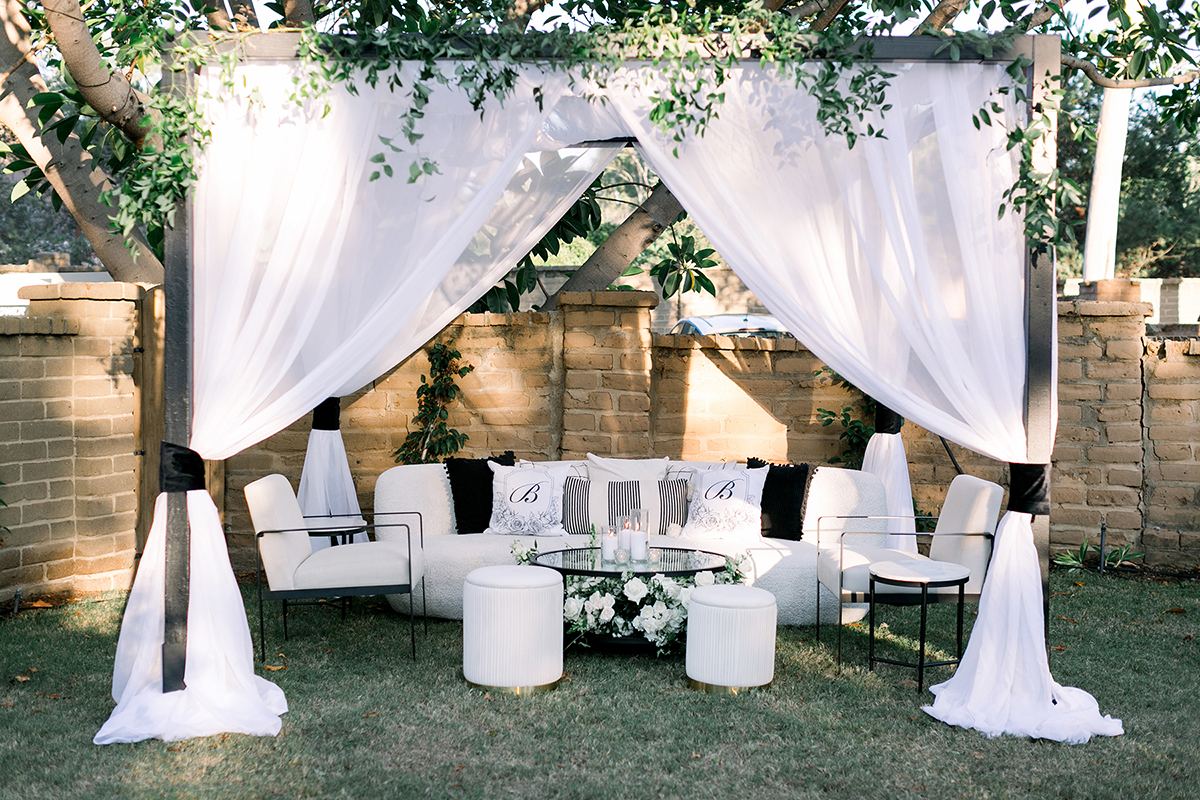 A reception lounge draped in white fabric, with white seating, monogrammed pillows, and a glass cocktail table.