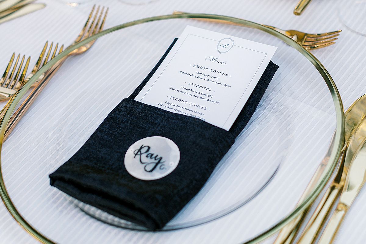A place setting with a gold-rimmed glass charger, gold flatware, a black napkin, and a printed menu.