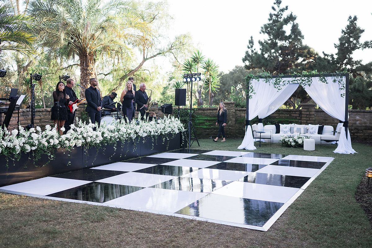 A black-and-white checkered dance floor set before the band, with the white-draped lounge and palms beyond.