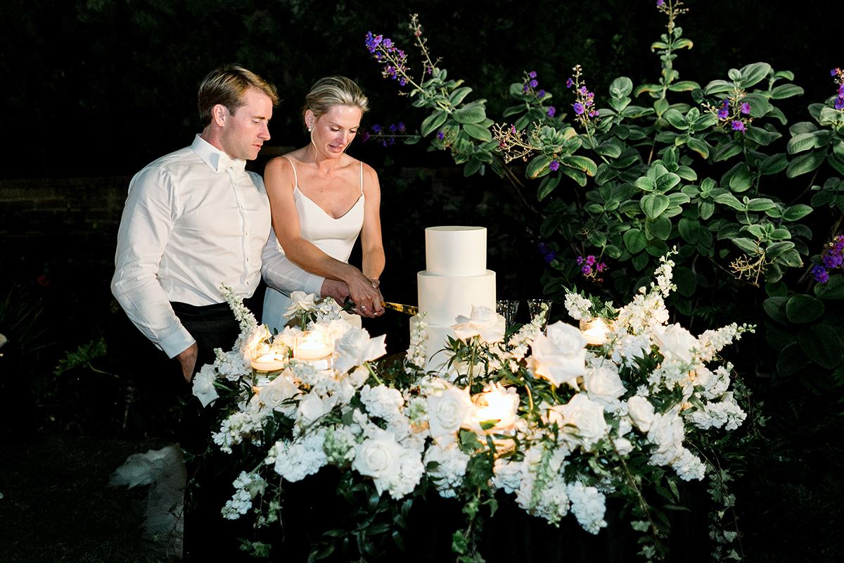 Claire and Collin cutting their wedding cake by candlelight, surrounded by white florals.