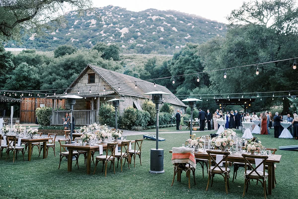 Outdoor vineyard-style wedding reception with rustic stone architecture, wood tables, and surrounding hills.