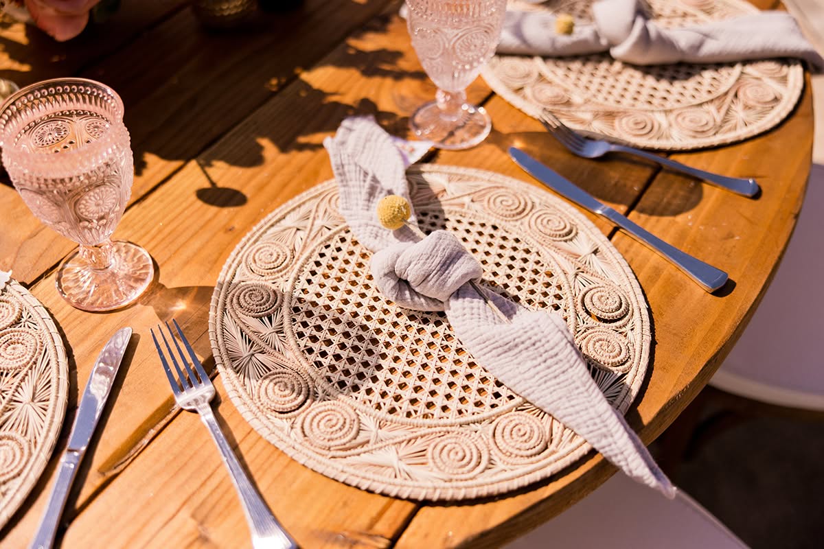 Outdoor wedding place setting with woven charger, patterned plate, blue flatware, and linen napkin.