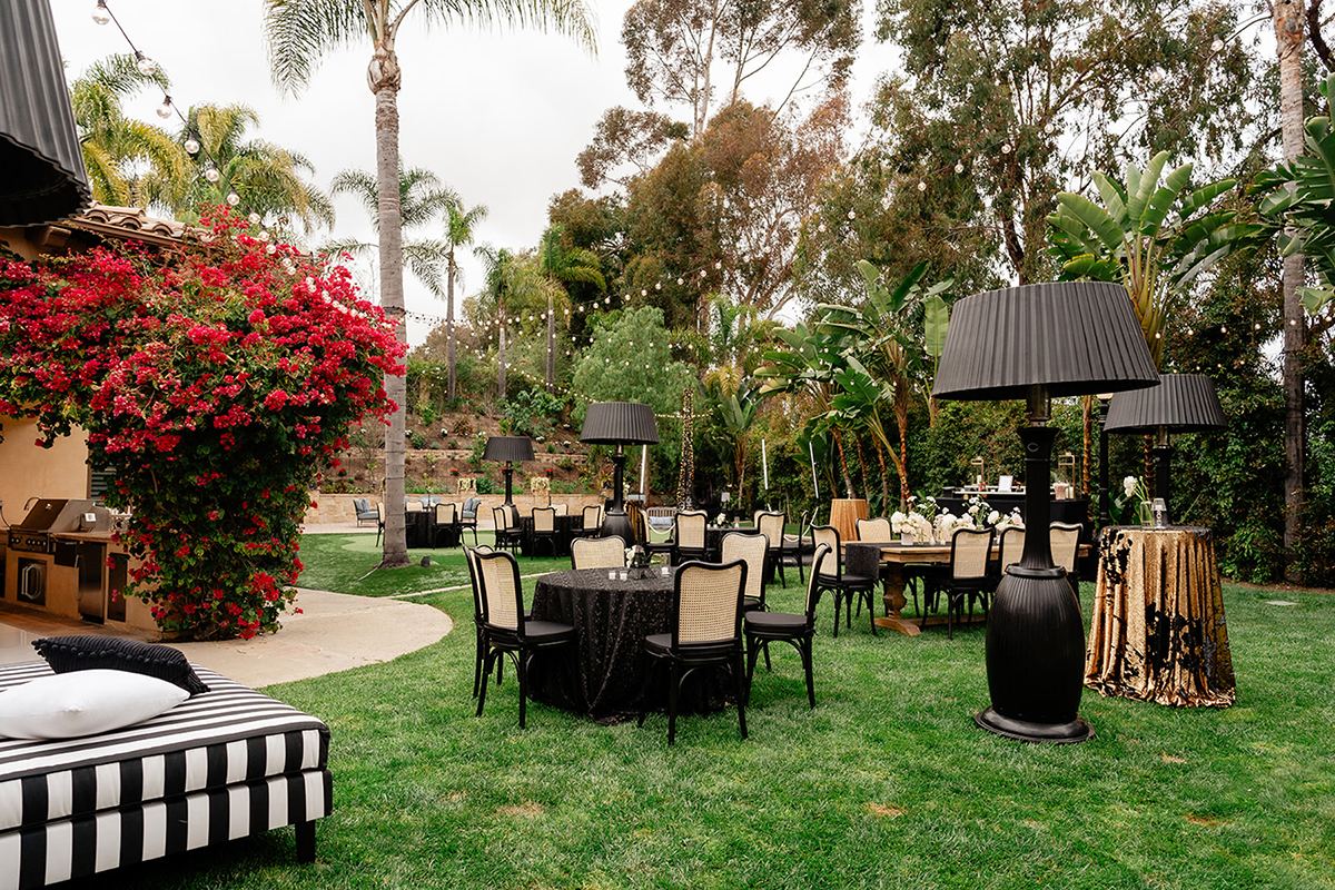 Outdoor garden party setup with black-and-white lounge seating, dining tables, black lamps, and palms.