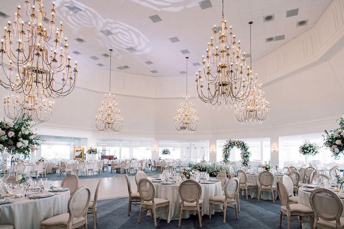 Elegant ballroom reception with chandeliers, white tables, and soft floral arrangements.