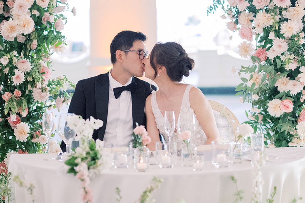 The bride and groom seated at their sweetheart table, framed by a lush pink and white floral arch.
