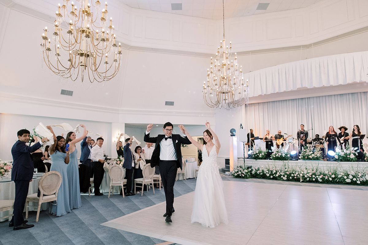 Bride and groom entering their reception beneath the ballroom chandeliers as guests cheer.