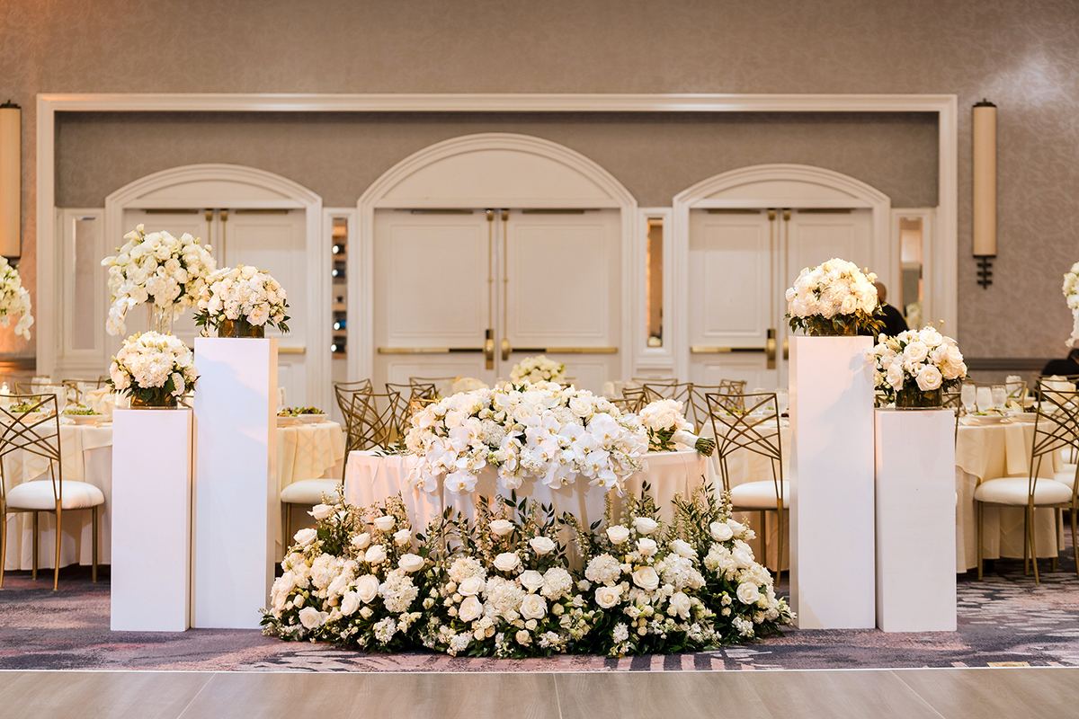 Reception sweetheart table with lush white florals and tall white pedestal arrangements in the ballroom.
