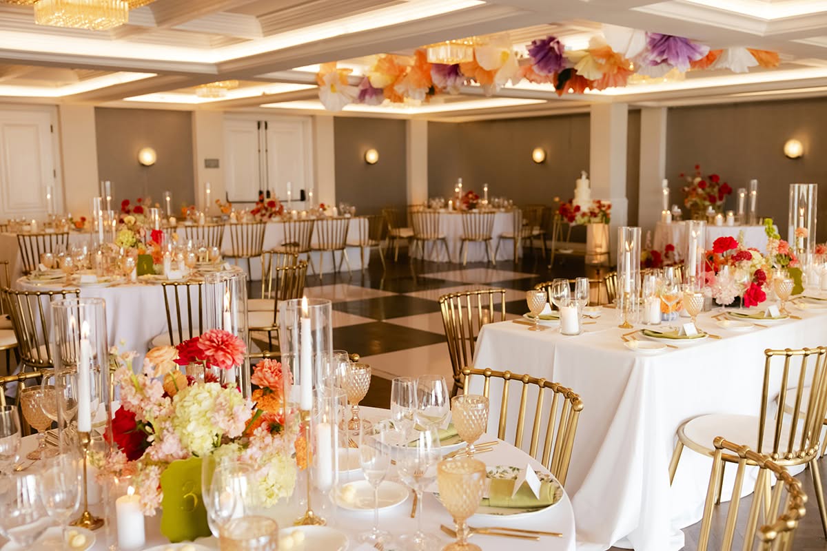 Reception ballroom with round tables, gold chairs, colorful floral centerpieces, and overhead décor.