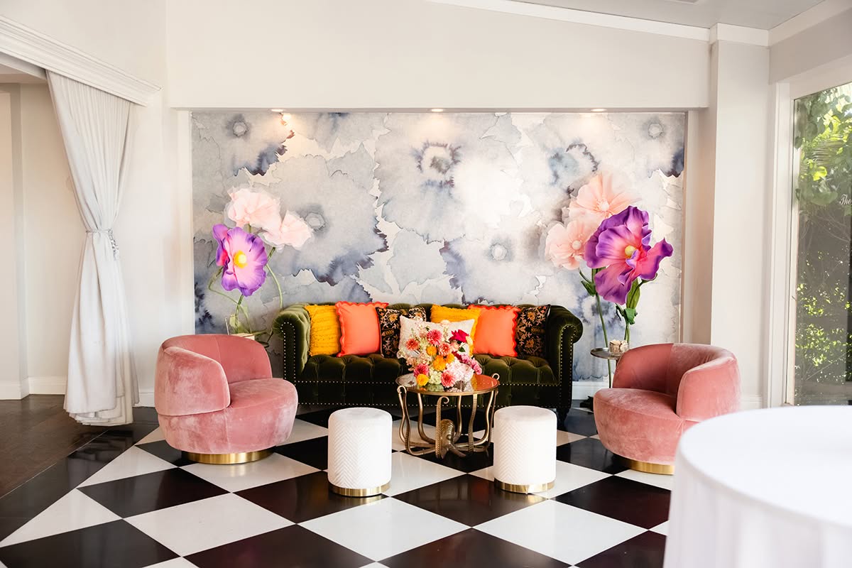 Lounge vignette with velvet seating, patterned pillows, bright florals, and a painted backdrop.