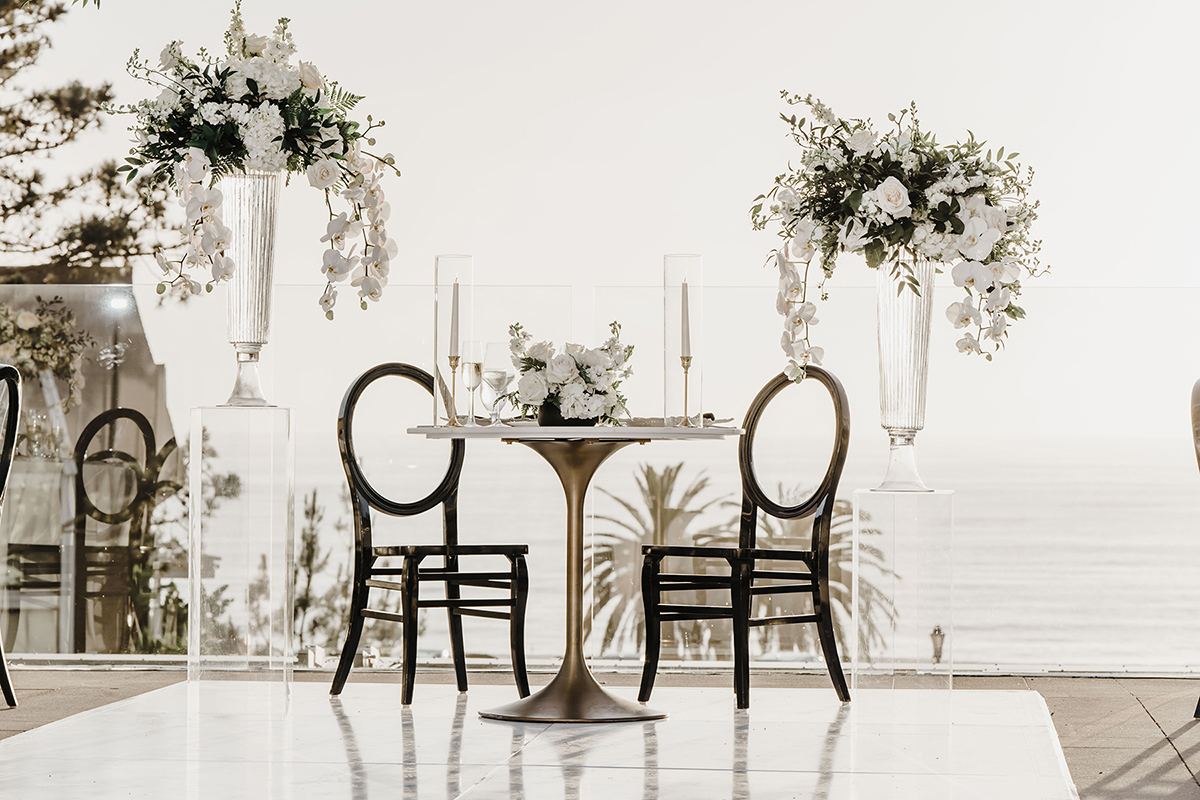 Sweetheart table with two black chairs, white florals, and tall floral arrangements set against an ocean view.