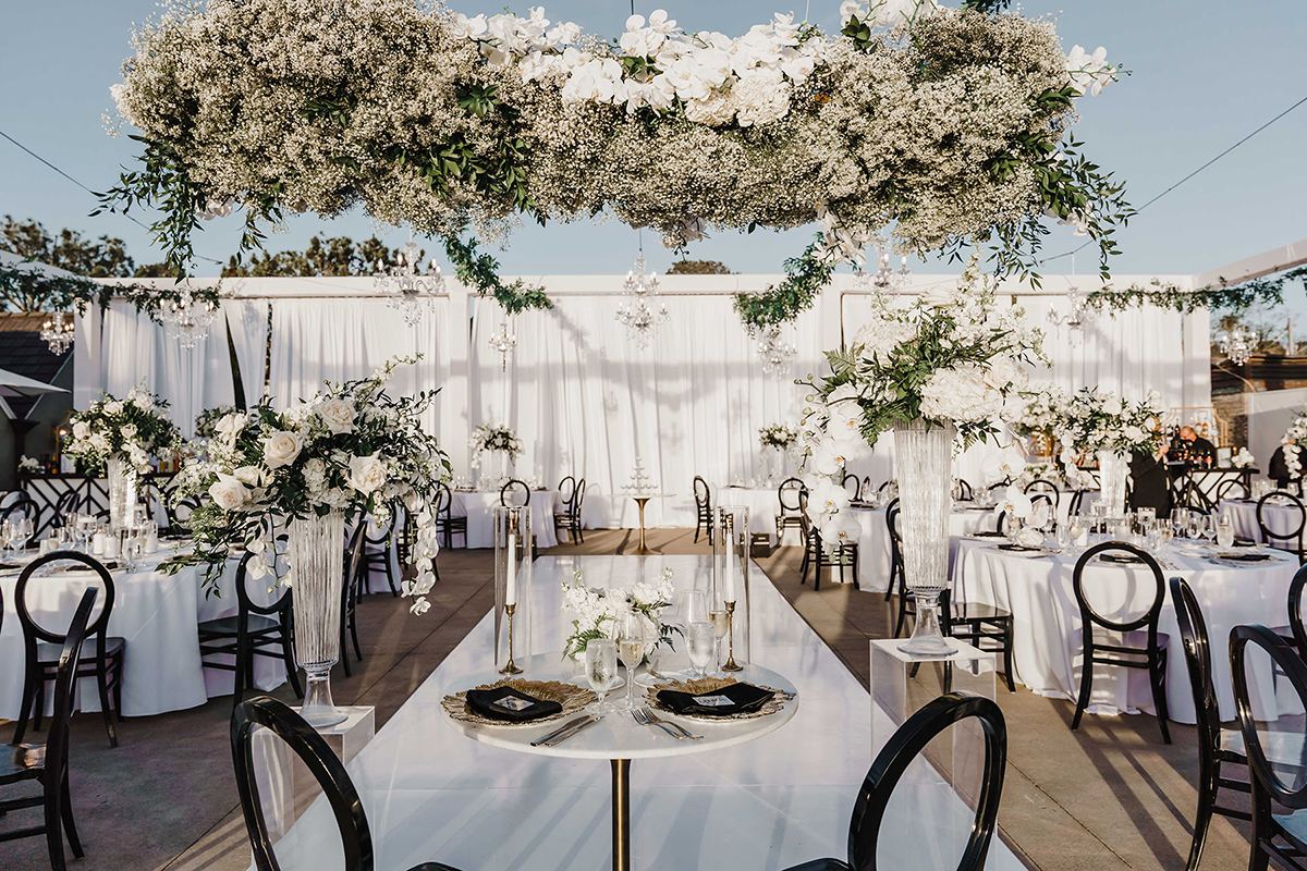 Reception table with black chairs, white florals, and an overhead floral installation at L’Auberge Del Mar.